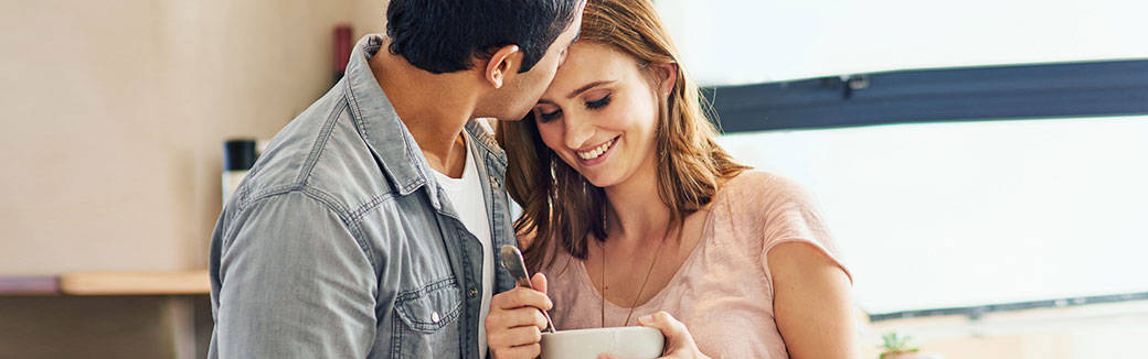 husband kissing the forehead of his wife, who is smiling & holding a cup of tea.